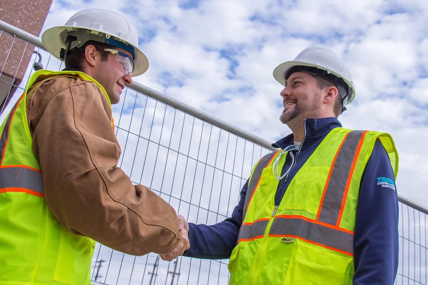 two construction workers in hard hats and safety vests shaking hands on a jobsite.