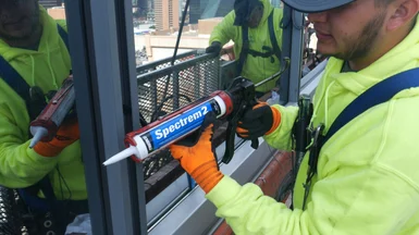 A worker is shown using a tube of Tremco Spectrem 2 silicone sealant in a caulk gun to apply glazing to an exterior window assembly.