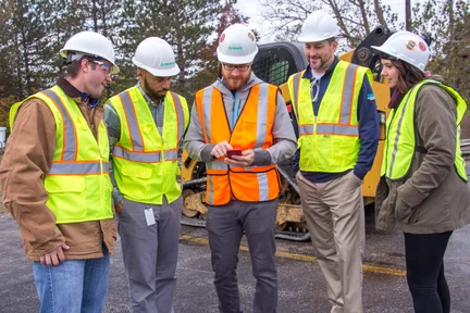 A group of construction workers observe while the site supervisor completes quality checks on a mobile device.