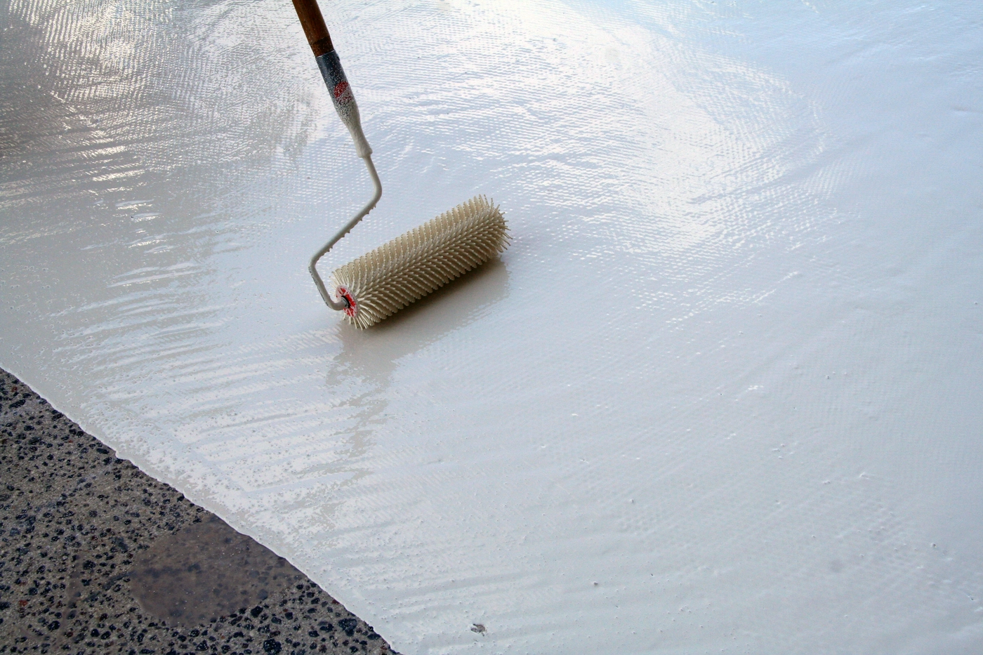 The picture depicts a spiked roller applying Tremco PUMA waterproofing to a floor. 